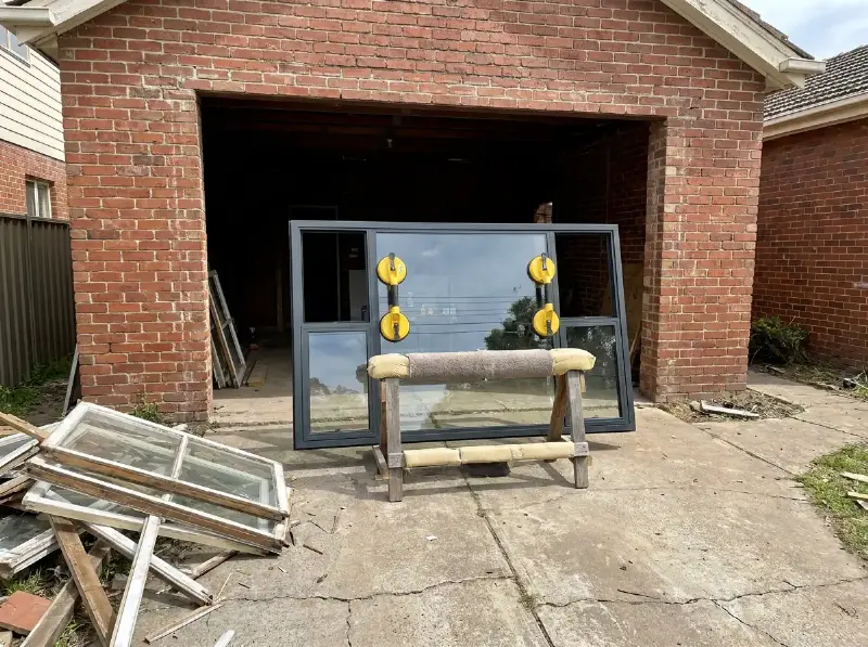 Nieuw aluminium raam op zuignappen en een steun klaar voor plaatsing in een open raamopening, oude ramen op de oprit